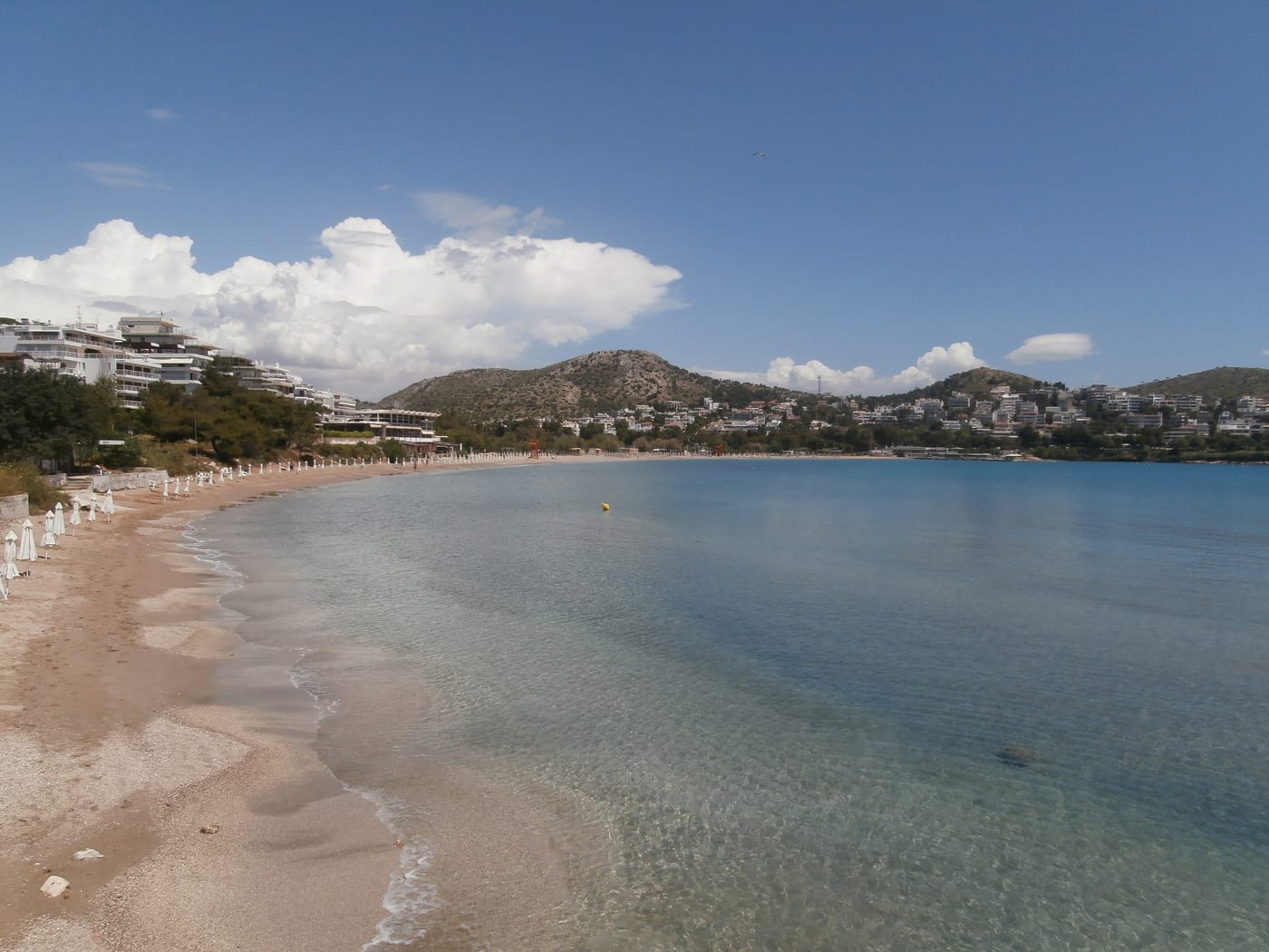 The Vouliagmeni shoreline on the Athens Riviera. Calm Aegean water and the curve of the bay. The prime residential stretch of the Athens coast.